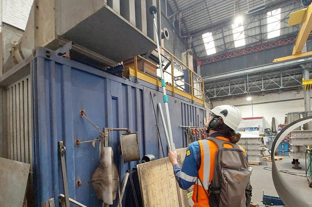 Acousticien équipé de ses EPI mesurant le bruit d'un équipement industriel à l'aide d'un microphone sur perche télescopique, lors d'une étude d'atelier